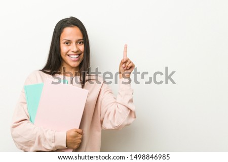 Young hispanic woman holding some notebooks smiling cheerfully pointing with forefinger away.