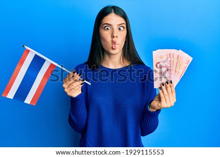 Young hispanic woman holding thailand flag and baht banknotes making fish face with mouth and squinting eyes, crazy and comical. 