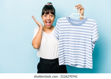 Young hispanic woman holding a t-shirt on a hanger isolated on blue background receiving a pleasant surprise, excited and raising hands. - Powered by Shutterstock