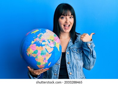 Young Hispanic Woman Holding World Ball Pointing Thumb Up To The Side Smiling Happy With Open Mouth 