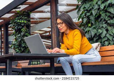 Young, Hispanic Woman With Indigenous Features Wearing A Yellow Sweater Sitting On A Bench Outside While Using Her Laptop