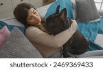 A young hispanic woman is indoors in her living room, cuddling with her pet french bulldog on a cozy sofa.