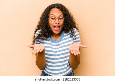 Young Hispanic Woman Isolated On Beige Background Holding Something With Palms, Offering To Camera.