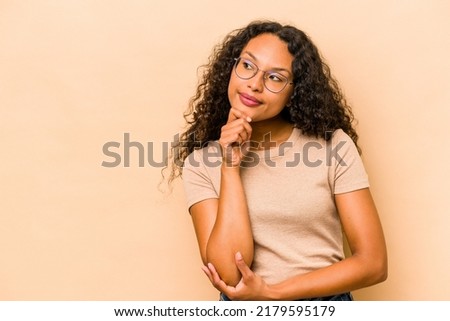 Young hispanic woman isolated on beige background looking sideways with doubtful and skeptical expression.