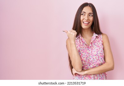 Young Hispanic Woman With Long Hair Standing Over Pink Background Smiling With Happy Face Looking And Pointing To The Side With Thumb Up. 