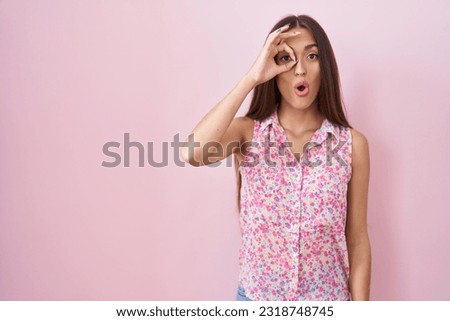 Young hispanic woman with long hair standing over pink background doing ok gesture shocked with surprised face, eye looking through fingers. unbelieving expression. 