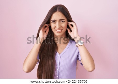 Young hispanic woman with long hair standing over pink background covering ears with fingers with annoyed expression for the noise of loud music. deaf concept. 