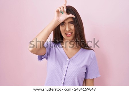 Young hispanic woman with long hair standing over pink background making fun of people with fingers on forehead doing loser gesture mocking and insulting. 