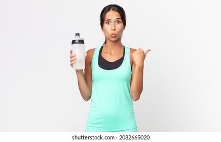 young hispanic woman looking astonished in disbelief and holding a water bottle. fitness concept