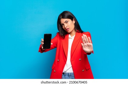 Young Hispanic Woman Looking Serious, Stern, Displeased And Angry Showing Open Palm Making Stop Gesture. Phone Screen Copy Space