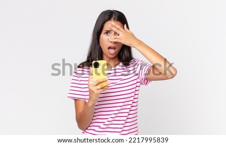 young hispanic woman looking shocked, scared or terrified, covering face with hand and holding a smartphone