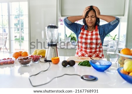 Young hispanic woman making healthy smoothie suffering from headache desperate and stressed because pain and migraine. hands on head. 