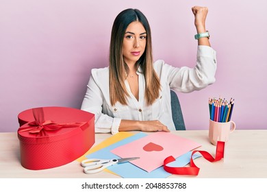 Young Hispanic Woman Making Valentine Gift Sitting On The Table Strong Person Showing Arm Muscle, Confident And Proud Of Power 