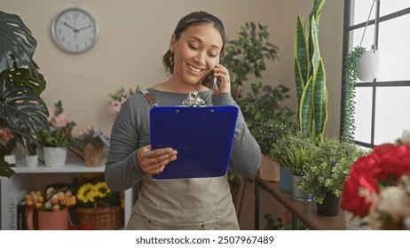 Young hispanic woman managing her flower shop, smiling while talking on the phone and holding a clipboard, surrounded by green plants and flowers. - Powered by Shutterstock