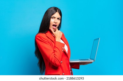 Young Hispanic Woman With Mouth And Eyes Wide Open And Hand On Chin, Feeling Unpleasantly Shocked, Saying What Or Wow. Laptop Concept