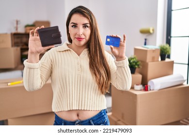 Young Hispanic Woman Moving To A New Home Holding Wallet And Credit Card Relaxed With Serious Expression On Face. Simple And Natural Looking At The Camera. 