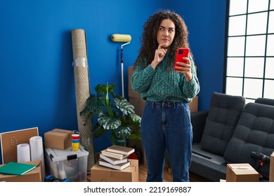 Young Hispanic Woman At New Home Using Smartphone Serious Face Thinking About Question With Hand On Chin, Thoughtful About Confusing Idea 