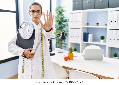 Young Hispanic Woman As Nutritionist Doctor Holding Weighing Machine With Open Hand Doing Stop Sign With Serious And Confident Expression, Defense Gesture 