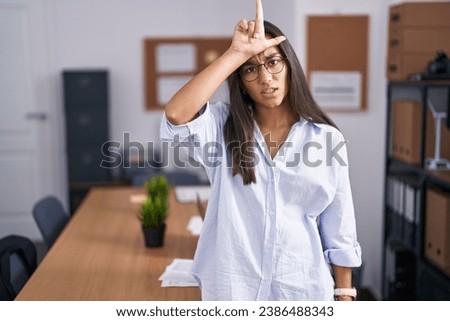 Young hispanic woman at the office making fun of people with fingers on forehead doing loser gesture mocking and insulting. 