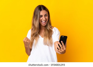 Junge hispanische Frau auf isoliertem gelbem Hintergrund mit Mobiltelefon und mit Siegesgestik