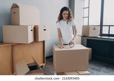 Young hispanic woman is packing containers with sticky tape. Happy mover is wrapping cardboard boxes with packing tape. Moving service worker preparing boxes for shipping and storage.