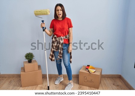 Young hispanic woman painting home walls with paint roller angry and mad screaming frustrated and furious, shouting with anger. rage and aggressive concept. 