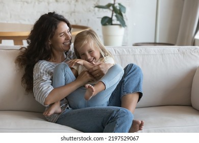 Young Hispanic Woman Play With Cute Little 5s Daughter, Having Playful Mood, Enjoy Playtime Seated On Cozy Couch At Home Looking Overjoyed Feeling Love. Happy Motherhood, Adoption, Family Bond Concept