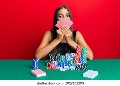 Young hispanic woman playing gambling poker covering face with cards relaxed with serious expression on face. simple and natural looking at the camera. 