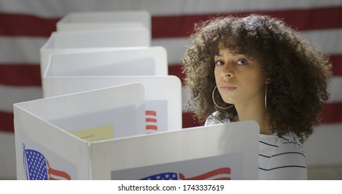  Young Hispanic Woman In Polling Station, Voting In A Booth With US Flag In Background. Serious Expression From High Viewpoint