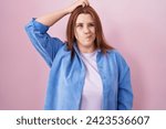 Young hispanic woman with red hair standing over pink background confuse and wondering about question. uncertain with doubt, thinking with hand on head. pensive concept. 