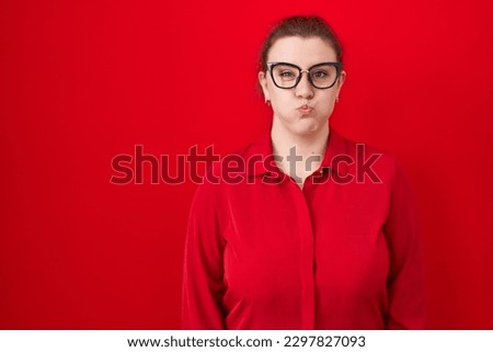Young hispanic woman with red hair standing over red background puffing cheeks with funny face. mouth inflated with air, crazy expression. 