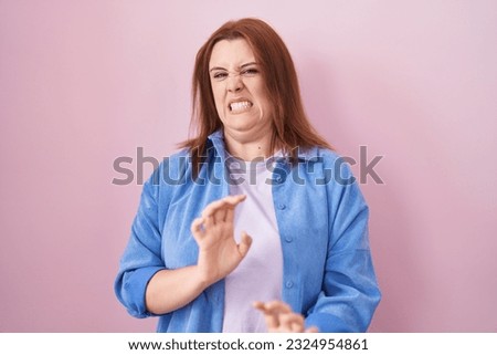 Young hispanic woman with red hair standing over pink background disgusted expression, displeased and fearful doing disgust face because aversion reaction. 