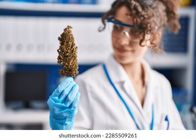 Young hispanic woman scientist smiling confident holding marihuana plant at laboratory
