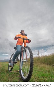 Young Hispanic Woman Seen From Below Wearing Orange Jacket And Blue Pants Riding Her Bicycle Alone Through The Rural Countryside During A Cloudy Day