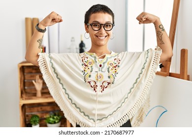 Young Hispanic Woman With Short Hair At Art Studio Showing Arms Muscles Smiling Proud. Fitness Concept. 