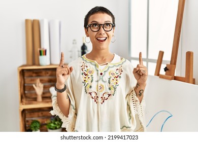 Young Hispanic Woman With Short Hair At Art Studio Amazed And Surprised Looking Up And Pointing With Fingers And Raised Arms. 