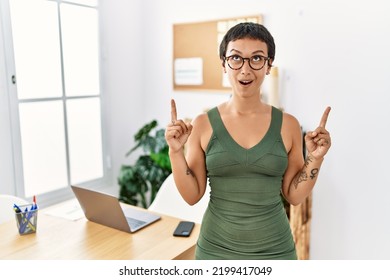 Young Hispanic Woman With Short Hair Working At The Office Amazed And Surprised Looking Up And Pointing With Fingers And Raised Arms. 