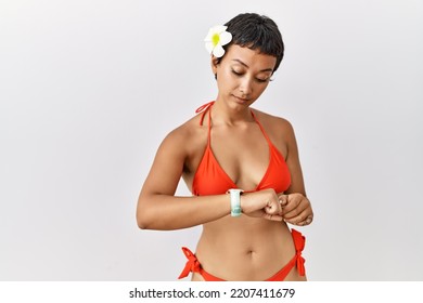 Young Hispanic Woman With Short Hair Wearing Bikini Checking The Time On Wrist Watch, Relaxed And Confident 