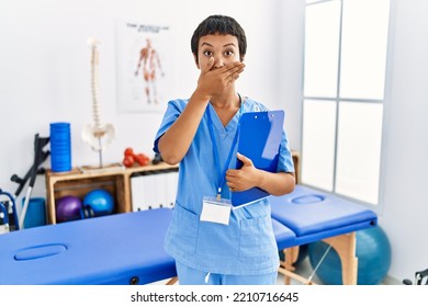 Young Hispanic Woman With Short Hair Working At Pain Recovery Clinic Shocked Covering Mouth With Hands For Mistake. Secret Concept. 