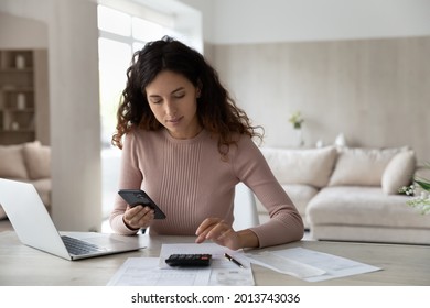 Young Hispanic woman sit at desk at home manage household finances paying bills calculating on machine cellphone. Millennial Latin female care of budget use smartphone for payment. Finance concept.