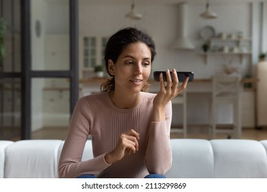 Young Hispanic woman sit on sofa holds smartphone near ear listens audio message, enjoy conversation on speaker phone. Sharing voicemail, remote communication, modern tech easy comfort usage concept