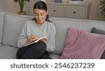 A young hispanic woman sits comfortably on a living room couch while writing in a notebook, in an inviting indoor home setting featuring a cozy atmosphere and attractive decor.