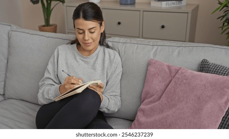 A young hispanic woman sits comfortably on a living room couch while writing in a notebook, in an inviting indoor home setting featuring a cozy atmosphere and attractive decor. - Powered by Shutterstock