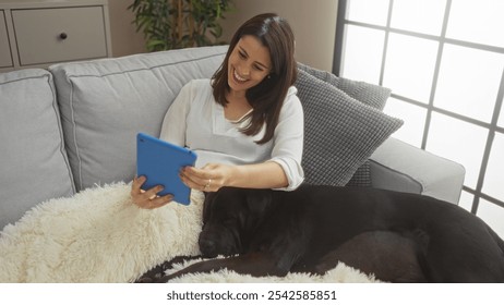 A young hispanic woman sits on a couch in an urban setting, smiling while looking at her tablet with her black labrador dog resting beside her. - Powered by Shutterstock