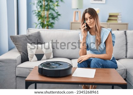 Young hispanic woman sitting at home by vacuum robot speaking on the phone thinking attitude and sober expression looking self confident  Foto stock © 