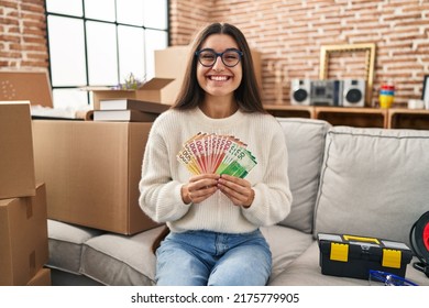 Young Hispanic Woman Sitting On The Sofa At New Home Holding Money Celebrating Crazy And Amazed For Success With Open Eyes Screaming Excited. 