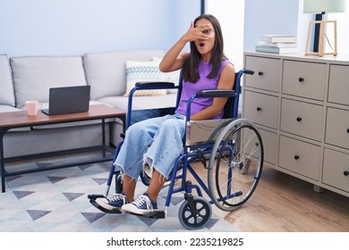 Young hispanic woman sitting on wheelchair at home peeking in shock covering face and eyes with hand, looking through fingers with embarrassed expression.  - Powered by Shutterstock