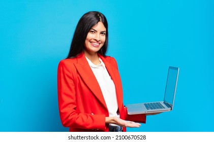 young hispanic woman smiling cheerfully, feeling happy and showing a concept in copy space with palm of hand. laptop concept