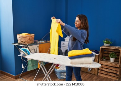 Young Hispanic Woman Smiling Confident Holding Clothes At Laundry Room