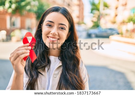 Young hispanic woman smiling happy holding red hiv ribbon walking at the city.